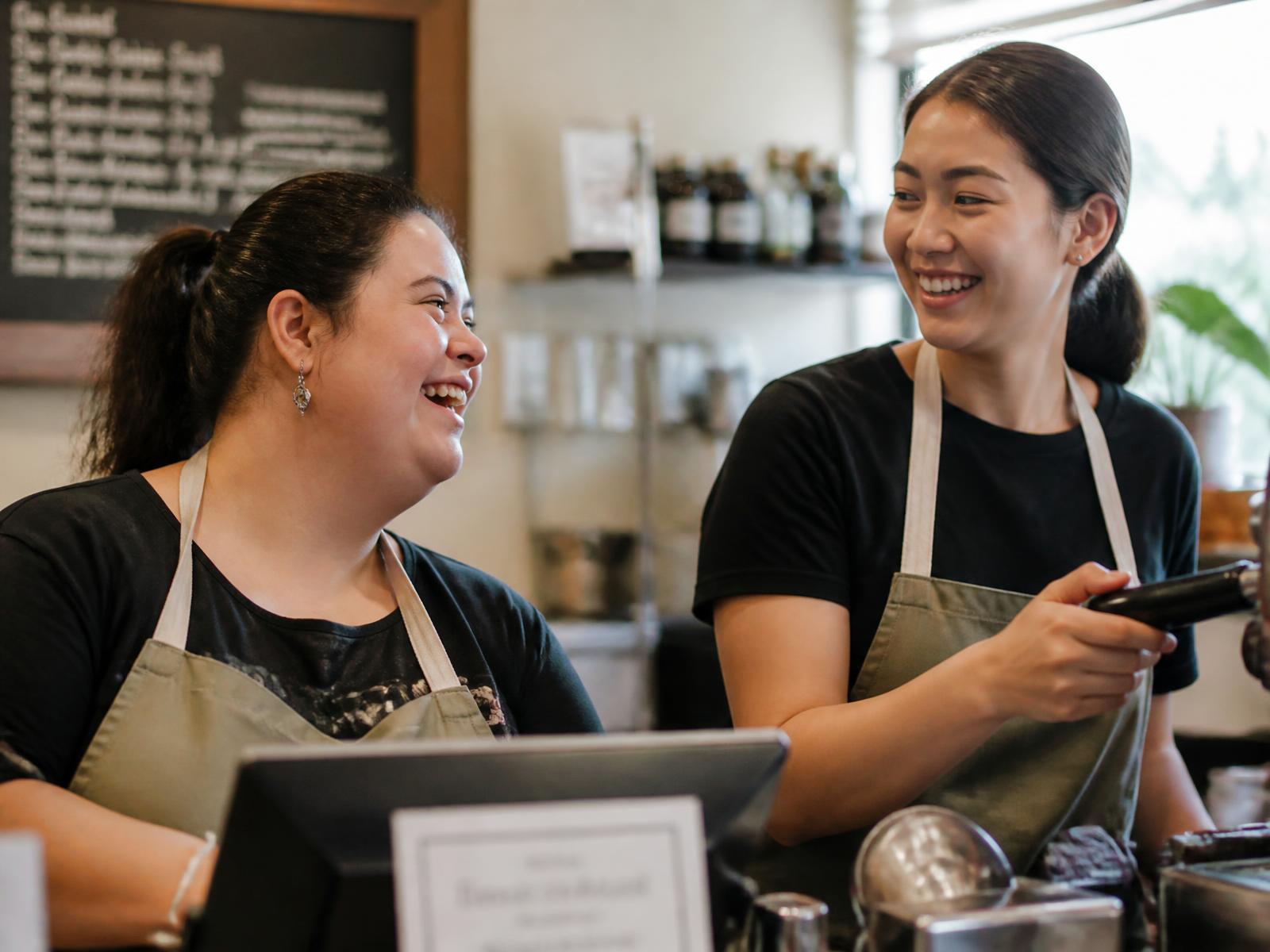 A woman working behind a neighborhood cafe counter, laughing with a coworker